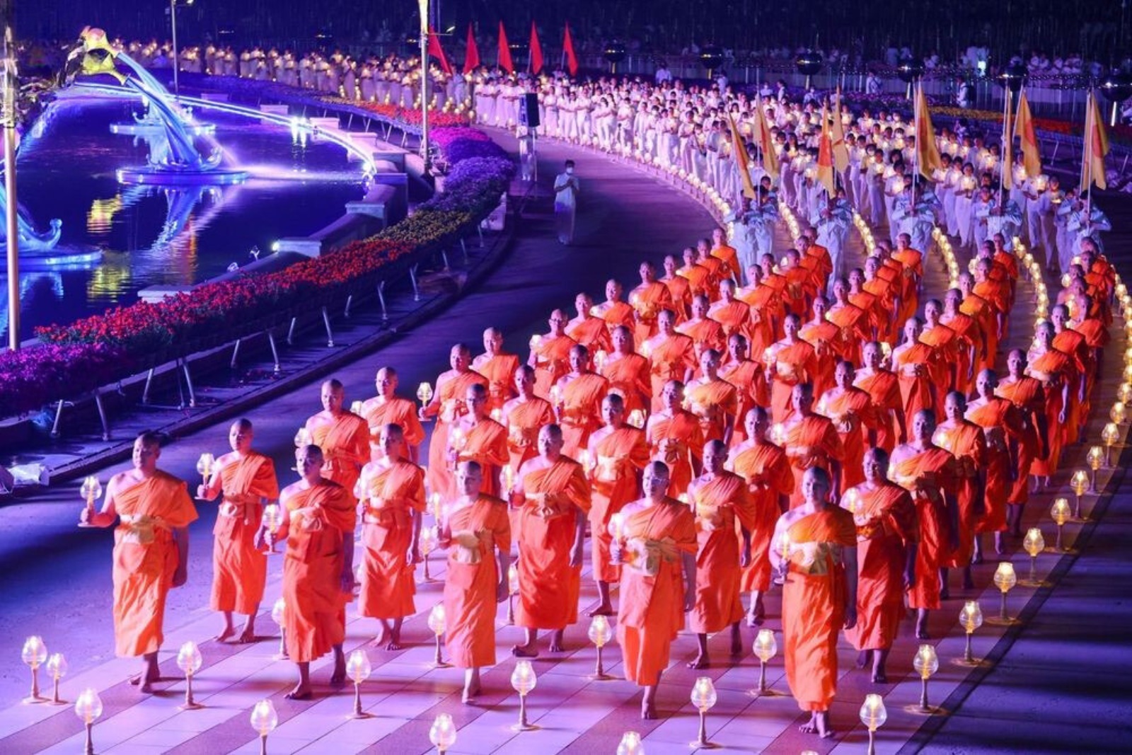 Ordained monks walking in file at night, each carrying a lit lamp, past the fountains at the temple in Thailand